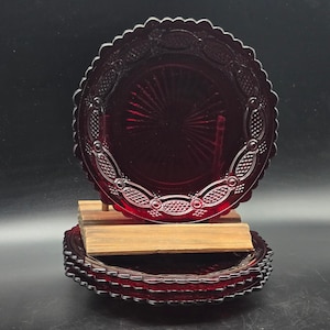 May include: A set of four circular ruby red glass plates featuring a scalloped edge and a decorative embossed border pattern of interconnected ovals and textured diamonds, with a central sunburst design radiating from the middle of each plate.