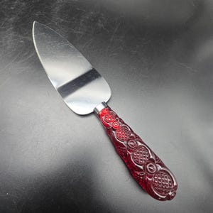 May include: A cake server featuring a polished stainless steel triangular blade with a serrated edge and a deep red, translucent acrylic handle. The handle displays a detailed, molded geometric pattern consisting of cross-hatched grids and circles.