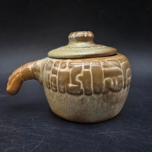 May include: A brown ceramic lidded pot with a handle. The pot has a textured surface with a repeating pattern. The lid has a small knob. The handle is curved and extends from the side of the pot.