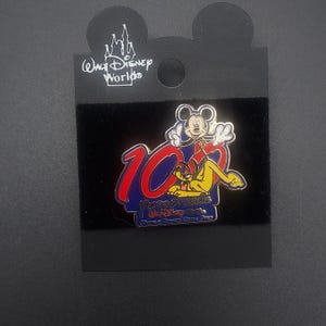 May include: A gold-colored pin featuring Mickey Mouse and Pluto celebrating 10 years of magic at Walt Disney World. The pin has a blue background with the text "Years of Magic" and "Walt Disney World" in white. The pin also includes the text "Where a Dream Comes True" in smaller white text.