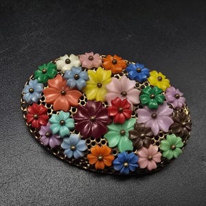 May include: An oval-shaped brooch featuring a gold-tone honeycomb metal base adorned with numerous small, multi-colored plastic flowers. The floral arrangement includes shades of red, orange, yellow, green, blue, purple, pink, and brown.