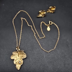 May include: A gold-toned jewelry set featuring a leaf pendant necklace and matching clip-on earrings. The necklace has a delicate chain and a detailed leaf pendant with a small, clear stone. The earrings are also leaf-shaped.
