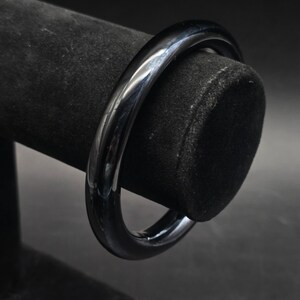 May include: A glossy, black bangle bracelet. The bracelet is a solid, circular band with a smooth, reflective surface. The jewelry is displayed on a black velvet jewelry stand.