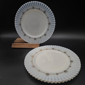 May include: Two white milk glass plates with scalloped edges. The upright plate features a blue-tinted rim and a black geometric floral pattern. The stacked plates below have a gold-toned geometric floral pattern along the scalloped border.