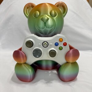 May include: A 3D-printed rainbow-colored teddy bear figure holding a white Xbox controller. The bear's head and limbs transition through shades of green, orange, and pink. The controller features colored buttons and a central Xbox logo.
