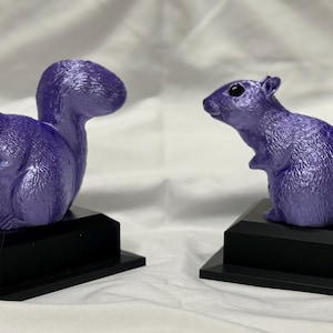 May include: Two purple squirrel figurines, each mounted on a black rectangular base. The squirrels have a glossy finish and are posed in a lifelike manner, with their tails arched. The background is a plain white cloth.