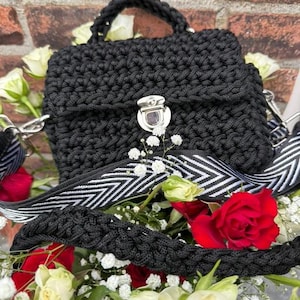 May include: A black crocheted handbag with a silver clasp and a black and white striped strap. The bag is surrounded by white and red roses.
