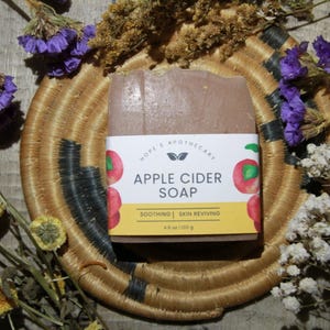 May include: A brown bar of apple cider soap with a white label that reads "Hope's Apothecary Apple Cider Soap Soothing | Skin Reviving 4.5 oz | 128 g" and features illustrations of apples.