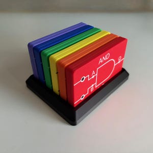 May include: A set of colorful circuit board coasters in a rainbow spectrum, displayed in a black holder. The coasters are red, orange, yellow, green, blue, and purple. The red coaster has the word "AND" and a circuit diagram.