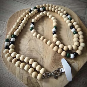 Könnte beinhalten: Ein Holzperlen-Lanyard mit silberfarbenem Verschluss. Das Lanyard hat naturfarbene Holzperlen, die mit schwarzen, weißen und grünen Perlen durchsetzt sind. Es wird auf einem Holzschneidebrett präsentiert.