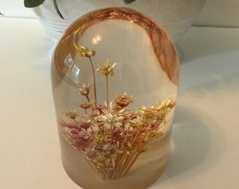 May include: A decorative resin paperweight featuring dried flowers encased within. The clear dome has a warm, amber-toned swirl design. The base is a soft, frosted beige. The flowers are a mix of yellow, white, and pink.