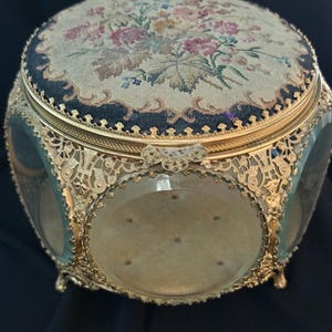 May include: An antique jewelry box with a floral tapestry lid and gold-toned filigree metalwork. The box has clear glass sides and a velvet interior, resting on small feet. The lid features a detailed needlepoint design with various flowers.
