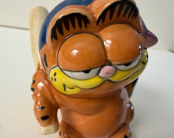 May include: Orange ceramic Garfield figurine with a blue hat and a yellow face. The figurine has black accents and a pink nose. A light-colored object is behind the figurine.
