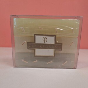 May include: A clear plastic box containing three rectangular bars of soap stacked vertically. The bars are light green, off-white, and sage green. A rectangular label in the center features the text CLAIRE BURKE and a stylized CB logo.