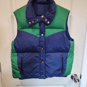 May include: A vintage, quilted vest in a color-block design. The vest features a navy blue center panel, green side panels, and a navy blue collar. It has a snap closure and two zippered pockets.