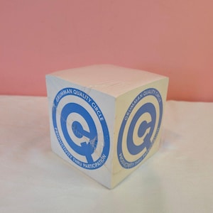 May include: A white cube-shaped notepad with blue text and a circular logo that reads "Grumman Quality Circle Productivity Thru Participation." The notepad is wrapped in clear plastic and sits on a white surface.