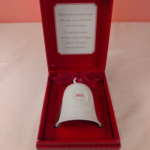 May include: A white ceramic bell ornament featuring a textured wreath design and the year 2007 in red text. The bell is tied with a red ribbon and presented in a red box with a printed message: Bells have been an important part of the magic and joy of Christmas for hundreds of years. Our 2007 bell continues the holiday tradition of bringing happiness and ringing in the spirit of the season.