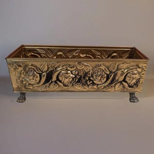 Puede incluir: Una jardinera rectangular de latón con un patrón en relieve de rosas florecientes y hojas en los lados exteriores. El recipiente se apoya sobre cuatro pequeñas patas detalladas en forma de garra en las esquinas, con acabado dorado.