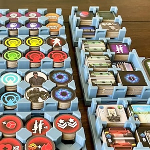 May include: A set of six blue plastic organizers with compartments for storing game pieces. The organizers contain various colored and patterned game pieces, including red, blue, green, yellow, and black. The organizers are arranged in a row on a wooden table.