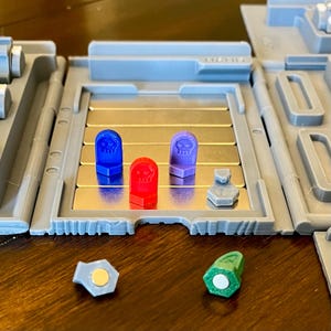 May include: A close-up of a game board with three small, colorful skull-shaped tokens. The tokens are red, blue, and purple. The board is made of metal and has a gray frame. There are also two small, gray hexagonal tokens on the board.