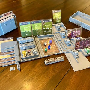 May include: A grey plastic organiser for a board game with compartments for cards, dice, and tokens. The organiser has the words "HEAL" and "DISCARD" printed on it.
