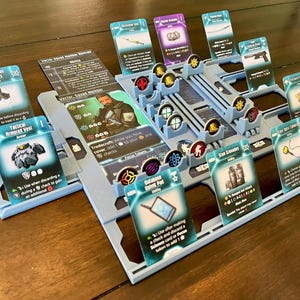 May include: A tabletop board game setup with various cards and game pieces. The cards feature illustrations of tactical gear, weapons, and character abilities. A light blue game board holds the cards and tokens, creating an organized play area.