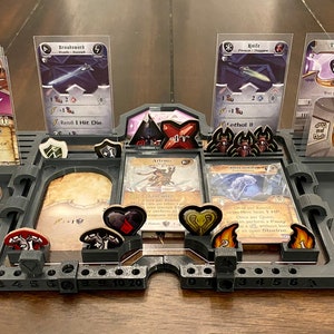 Può includere: Un organizzatore di gioco in plastica grigia con slot per carte, gettoni e dadi. L'organizzatore ha un design bianco e nero con la parola "Artemis" stampata su di esso. Sono visibili diverse carte, tra cui "Breakneck", "Knife", "Prophecy" e "1919".