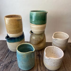 May include: A collection of handmade ceramic pitchers and cups in various colors and finishes. The pitchers have a spout for pouring. Colors include teal, green, tan, and speckled white. The pieces are arranged on a wooden surface.