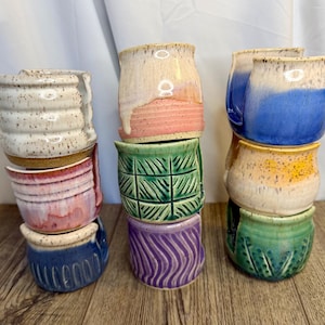 May include: Nine ceramic yarn bowls arranged in three vertical stacks. The bowls feature various glaze colors including white, pink, blue, green, purple, and yellow, with speckled textures and carved patterns like vertical lines, grids, and waves.