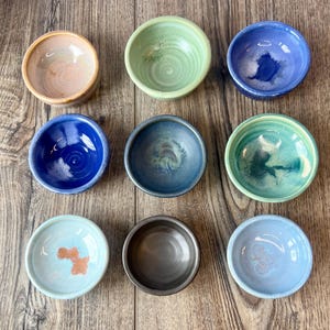 May include: Nine small, handmade ceramic bowls in various colors, including blue, green, and peach. Each bowl has a unique glaze and shape, perfect for serving dips or small portions. The bowls are arranged on a wooden surface.