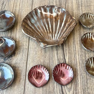 May include: A collection of ceramic serving dishes in various sizes and shapes. The set includes a large, shell-shaped bowl and several smaller, round and shell-shaped dishes. The colors range from iridescent bronze to reddish-pink, with a glossy finish.
