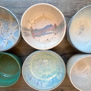 May include: Overhead shot of six ceramic bowls in various colors and designs. The bowls have a round shape and are glazed with colors including blue, green, and speckled white. The bowls are arranged on a wooden surface.