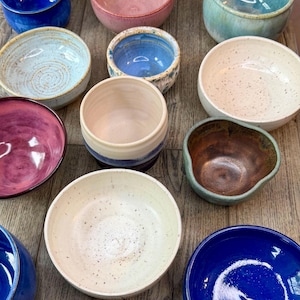 May include: A collection of handmade ceramic bowls in various colors and sizes. The bowls feature a speckled glaze in shades of blue, pink, green, and white. Some bowls have a smooth finish, while others have a textured appearance. The bowls are arranged on a wooden surface.