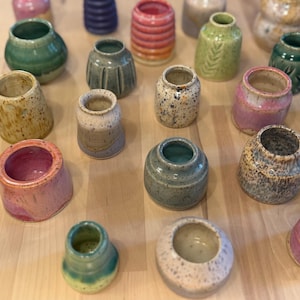 May include: A collection of small, handmade ceramic vases in various colors and textures. The vases are cylindrical and have a variety of finishes, including speckled, glazed, and textured surfaces. Colors include green, pink, blue, and yellow.
