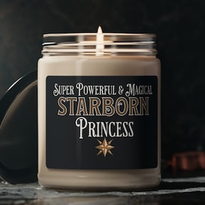 Puede incluir: Una vela beige en un tarro de vidrio con una etiqueta negra que dice "Super Powerful & Magical Starborn Princess" con una estrella dorada. La vela está encendida y la llama es visible.