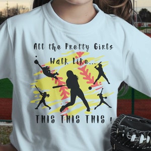 May include: A light blue t-shirt with a softball graphic and the text "All the Pretty Girls Walk Like... This This This!" The graphic features a softball player in silhouette with a red and yellow softball.