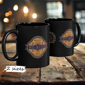 Puede incluir: Dos tazas de café negras con un logotipo de estilo vintage que dice "Two Rivers" en letras doradas. El logotipo está rodeado por un círculo dorado con un borde azul y rojo. El texto "It's Piping Good Stuff" está por encima del logotipo, y el texto "Smooth as the Winespring Water" está por debajo del logotipo.