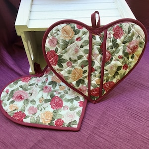 Puede incluir: Dos agarraderas de olla con forma de corazón acolchadas con un patrón floral en tonos de rosa, rojo, amarillo y verde. Las agarraderas tienen un ribete burdeos.