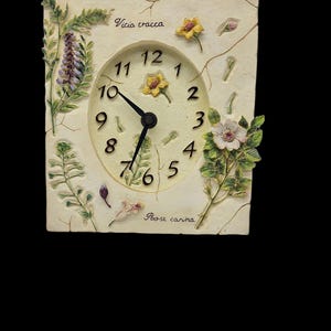 May include: A square, cream-coloured clock featuring a floral design. The clock face has black numerals and hands, with yellow and white flowers. The words "Vicia cracca" and "Rose carina" are written on the clock.