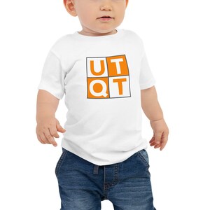 Pode incluir: Um macacão branco para bebê com mangas curtas que apresenta um quadrado laranja com as letras "UTQT" em branco.