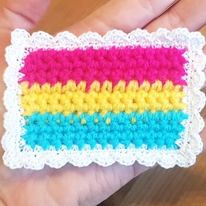 May include: A small crocheted square in the colours of the pansexual pride flag. The square is pink, yellow, and blue with a white crocheted border.
