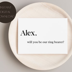 Op de afbeelding: Een witte kaart met de tekst "Alex, will you be our ring bearer?" is gecentreerd op een licht houten bord. Een donkerbruine cirkel met de woorden "INSTANT DIGITAL DOWNLOAD" staat in de linkerbovenhoek.