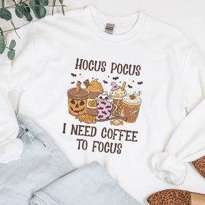May include: White sweatshirt with the text "Hocus Pocus I Need Coffee To Focus" and a graphic of Halloween-themed coffee drinks. The sweatshirt is paired with light wash jeans and leopard print flats.