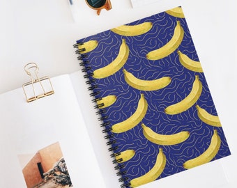 Puede incluir: Un cuaderno de espiral con un patrón de plátanos amarillos sobre un fondo azul oscuro con líneas onduladas doradas. El cuaderno está abierto, revelando páginas en blanco y una foto. Gafas de sol y un bolígrafo son visibles.