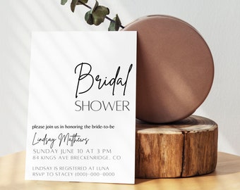 Puede incluir: Una tarjeta de invitación blanca con las palabras "Bridal Shower" en elegante escritura negra. La tarjeta incluye detalles sobre el evento, como la fecha, la hora y el lugar. Un jarrón marrón y un bloque de madera están en el fondo.