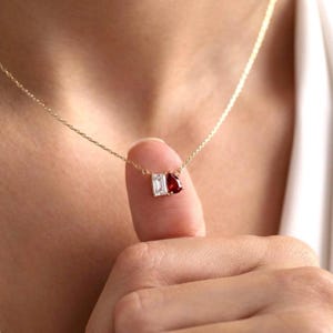 Peut inclure: Un collier à fine chaîne dorée orné de deux petites pierres précieuses suspendues au centre. L'une est une pierre transparente de taille émeraude rectangulaire, l'autre est une pierre rouge foncé en forme de poire, montées côte à côte.
