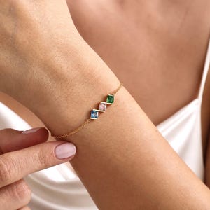 May include: A delicate gold chain bracelet featuring three square-cut gemstones arranged in a row. The stones are set in a diagonal pattern, showcasing a vibrant blue, a soft pink, and a deep green color, connected by thin gold metal bars.