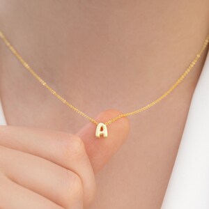 Könnte beinhalten: Eine zarte goldene Halskette mit einem Anhänger in Form des Buchstabens "A". Der Anhänger ist klein und an einer feinen Goldkette befestigt. Die Halskette wird nah am Hals getragen und erzeugt einen minimalistischen und eleganten Look. Die goldene Farbe sorgt für eine warme, klassische Ästhetik.