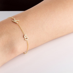 Puede incluir: Un delicado brazalete de cadena dorada con dos dijes de iniciales, uno con la letra "K" y el otro con la letra "S", ambos adornados con brillantes piedras de circonita cúbica.