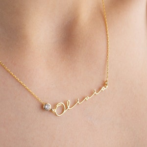 May include: A gold necklace with a delicate chain and a small diamond accent. The necklace features the name "Olivia" written in cursive.